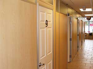 A corridor with numbered doors on the right side, featuring wooden walls and top panel glazing in a well-lit indoor space.