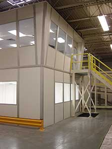 An industrial office module with large fixed windows, housed inside a warehouse, adjacent to a yellow metal staircase leading up to a second level.