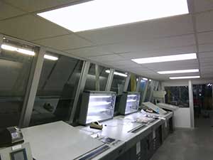 A dimly lit office with multiple computer monitors on desks, some turned on, under fluorescent ceiling lights. A tilted window adds a unique element to the space, which appears empty with no visible people.