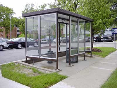 A glass-walled bus shelter on a sidewalk with seating, a trash can, and a vehicle parked on the street in the background. Green trees and grass surround the area, reminiscent of Windows updates providing