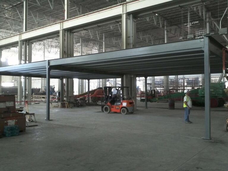 A spacious industrial warehouse with high ceilings, exposed steel beams, and integrated mezzanines. A worker in a high-visibility vest stands near a forklift carrying materials, with other machinery and