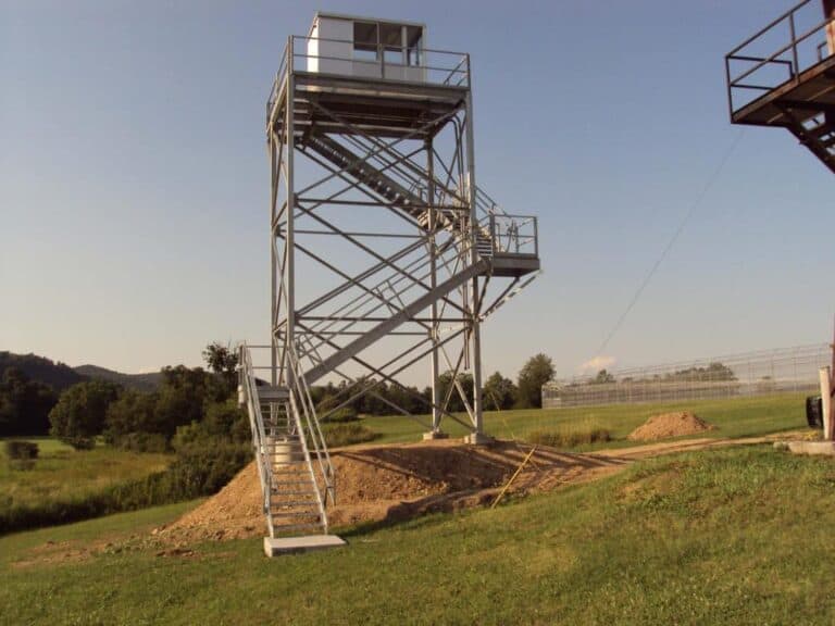 Metal observation tower with stairs in a grassy field under a clear sky, serving as a popular tourist attraction.