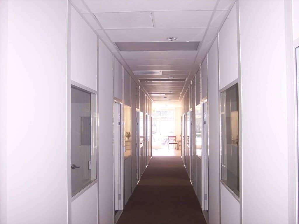 A long, brightly lit office corridor with glass doors on each side, carpet flooring, and a series of recessed ceiling lights leading towards a window at the end, flanked by modular office partitions.
