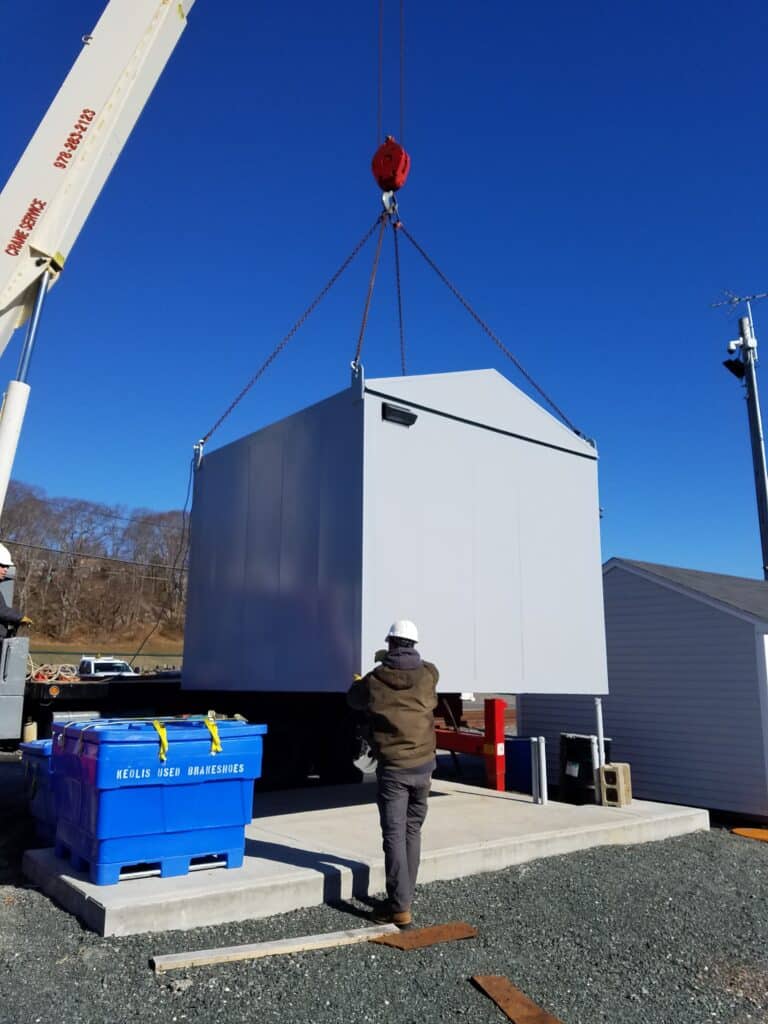 A worker in a hard hat oversees a large crane lifting a grey industrial container on a sunny day, with clear blue sky and other outdoor equipment enclosures nearby.