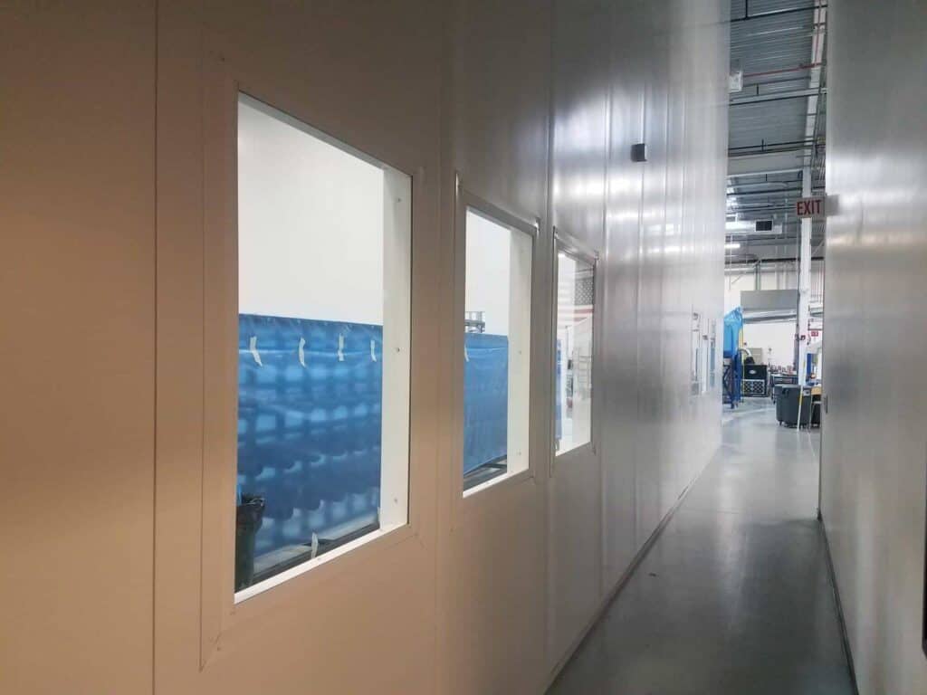 Tall Modular Wall in White 9 A hallway within an industrial or office space, featuring a row of windows along a white wall, overlooking a blue-curtained area, with activity and machinery visible in the background. The hallway is bordered