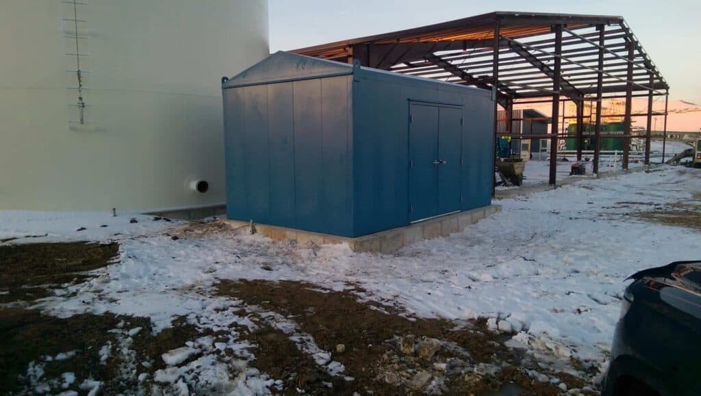 A blue industrial shed on a concrete base surrounded by snow, adjacent to a partially constructed metal framework and a large white tank, during early evening, is utilized as an outdoor equipment enclosure.