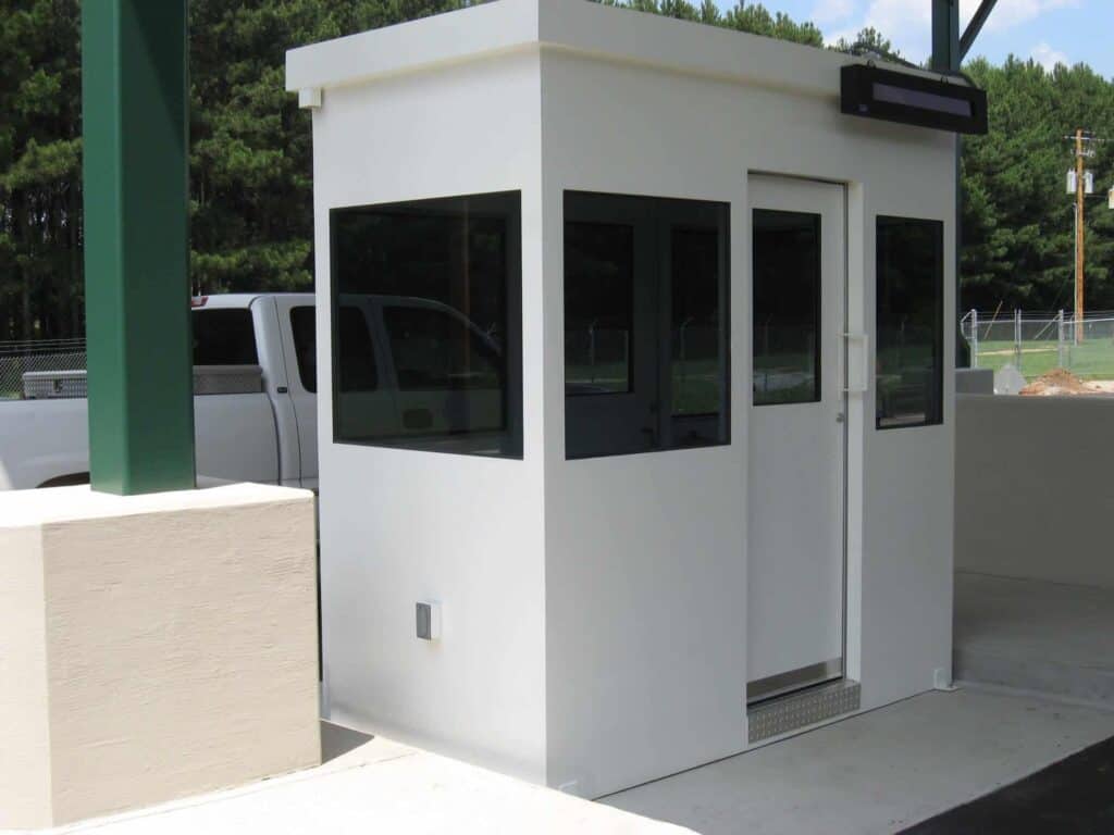 A small white security booth with multiple windows, located at the entrance of a parking area, under a clear blue sky.