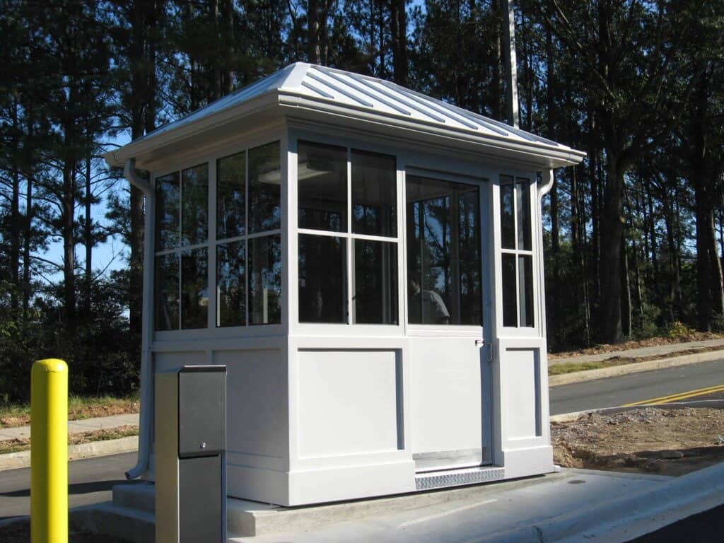 Guard/Security Booth 43 A small, white access control booth with large windows and a gray roof, located beside a road amid green trees under a clear blue sky.