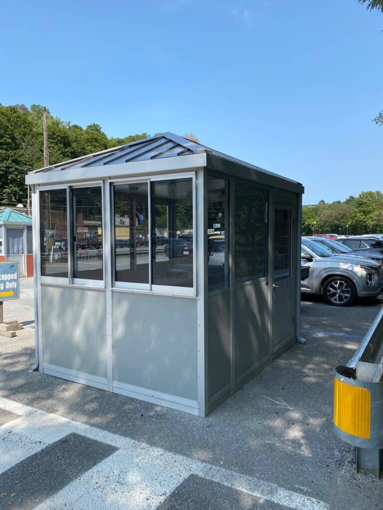 Guard/Security Booth 4 A small, gray guard booth with glass windows and a metal roof stands in a parking lot surrounded by cars under a clear blue sky.