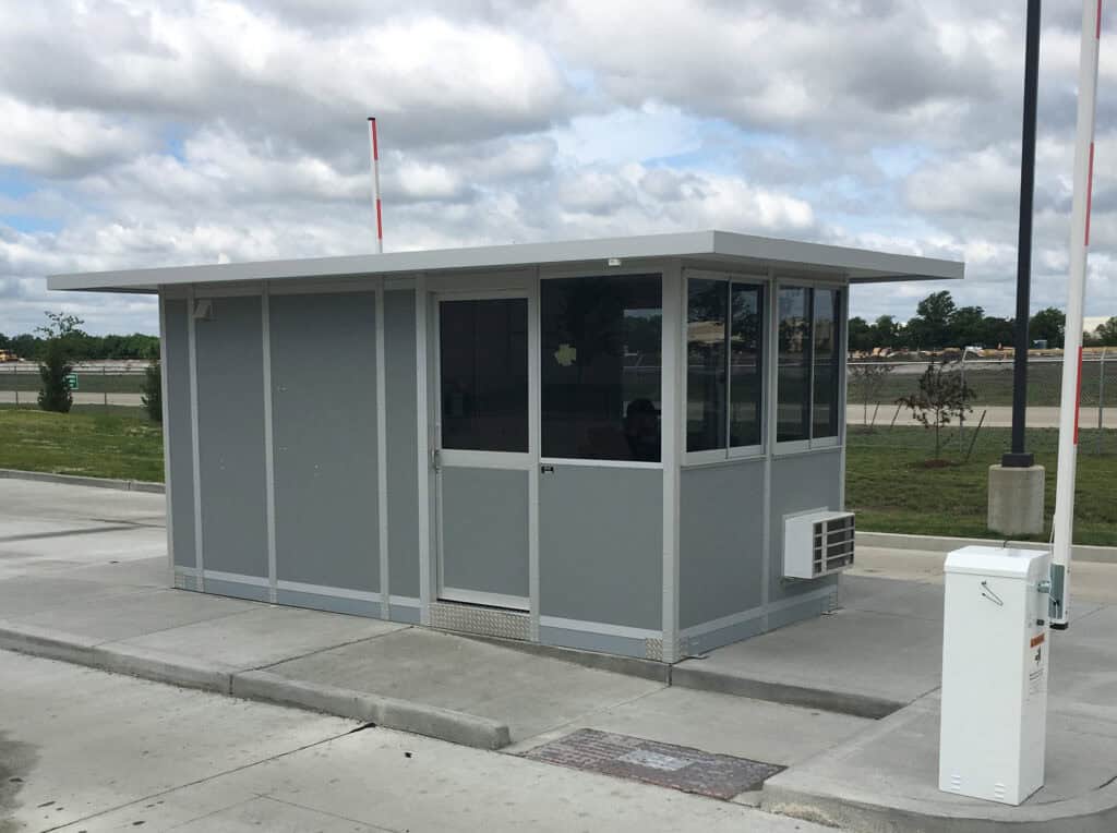 Guard/Security Booth 18 A small, modern security booth made of gray materials with large windows, situated on a concrete platform in an open area with cloudy skies overhead.