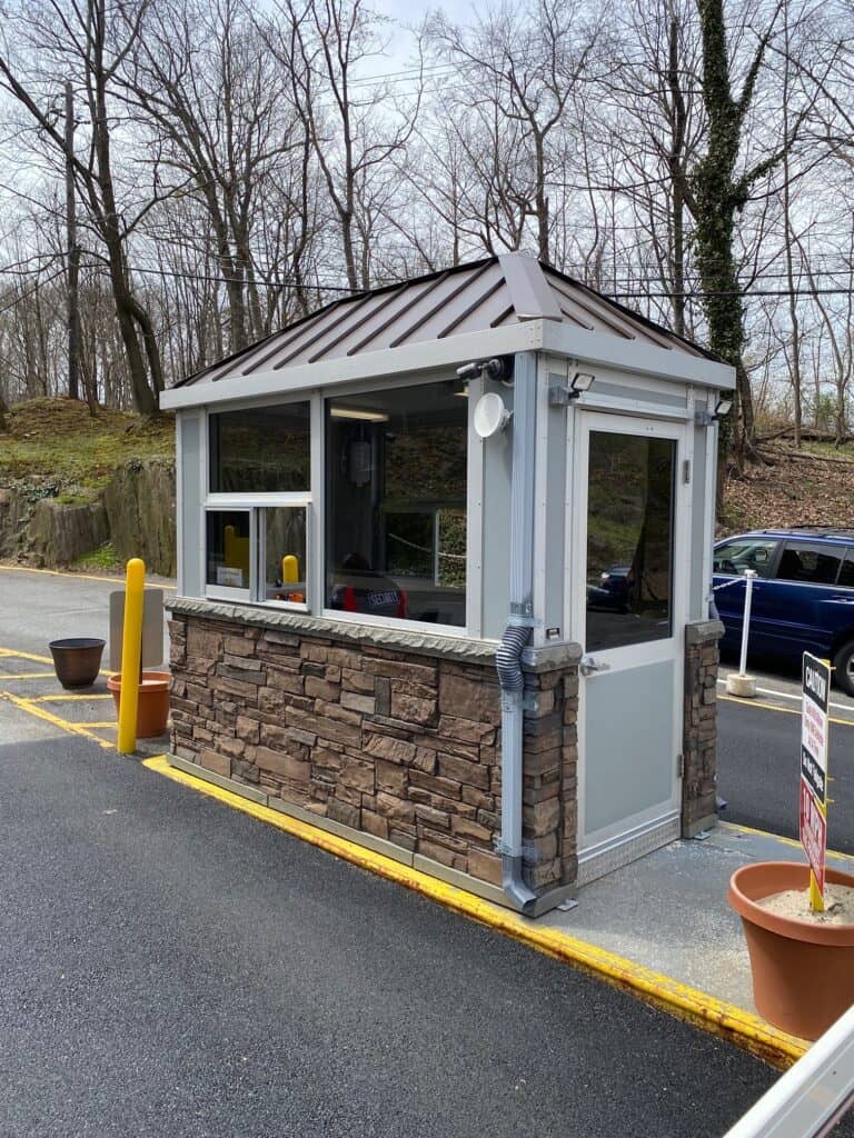 Guard/Security Booth 9 A small, well-maintained security booth constructed with a stone base and a gray metal frame, situated by a parking lot with scattered cars and leafless trees in the background.