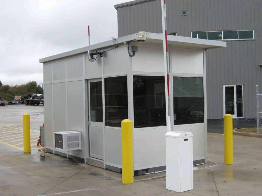 Guard/Security Booth 39 A small, modern security booth with glass windows and gray walls, located next to a large building. It features ventilation units, protective yellow bollards, and a white access control barrier.