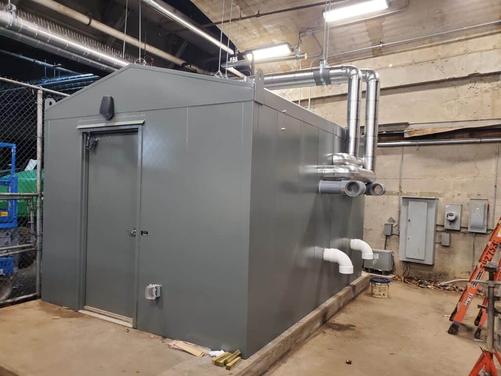 A large, gray industrial walk-in cooler with external piping in a utility room, featuring electrical enclosures on the right wall and a ladder leaning on the left.