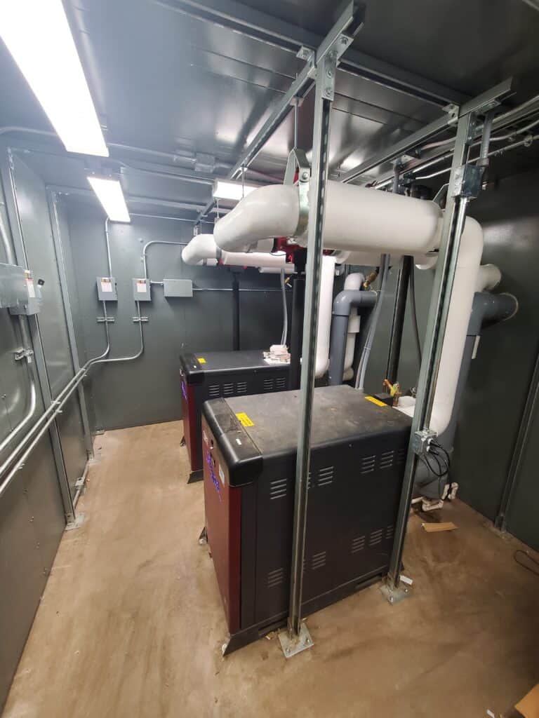 Industrial boiler room featuring a large black boiler, white pipes overhead, and a structural metal framework with equipment enclosures, set against grey walls lit by fluorescent lights.