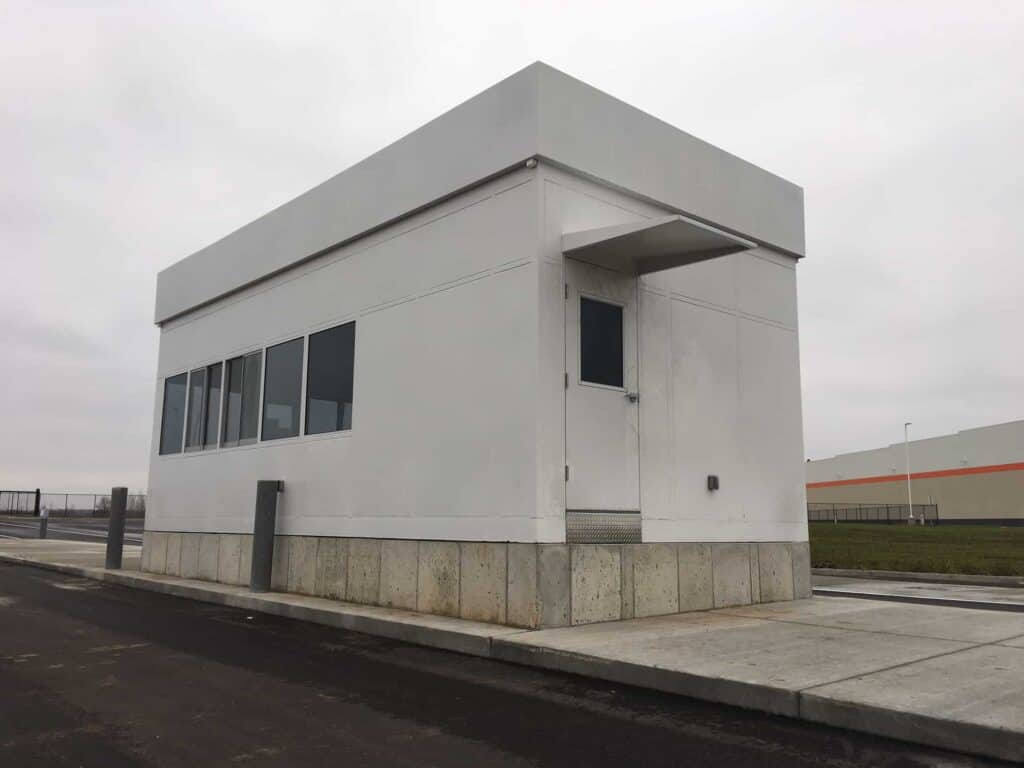 Guard/Security Booth 40 A modern, small white security booth with large windows, standing in a parking lot on an overcast day. The structure has a flat roof and is surrounded by a paved area.