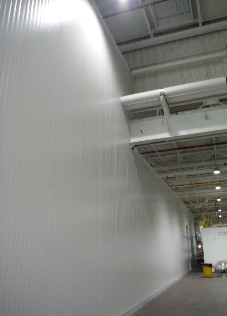 Tall Modular Walls Inside Auto Plant 11 Interior of an industrial building showing a long, curved tall wall with ribbed texture, under a ceiling with exposed metal beams and pipes. Fluorescent lights illuminate the area.