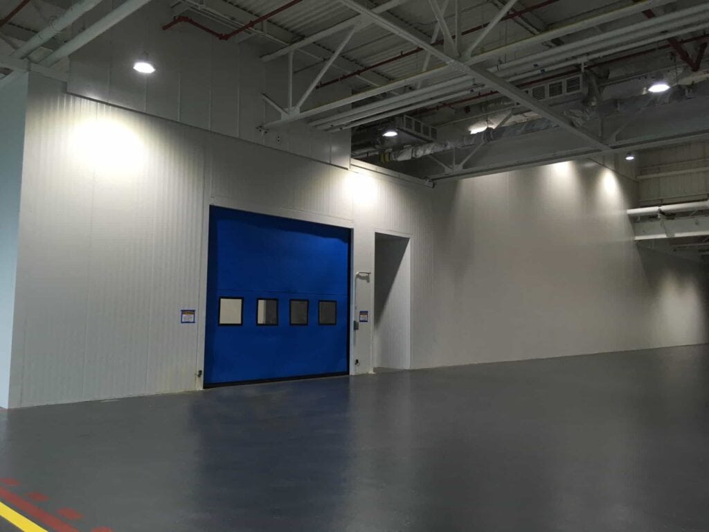 Tall Modular Wall in White 12 Large blue door with small windows in an industrial building, surrounded by tall walls and illuminated by ceiling lights, with a polished grey floor marked by yellow lines.