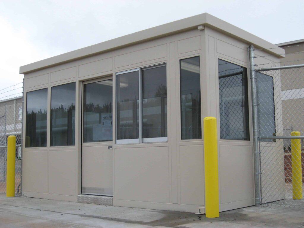 Guard/Security Booth 35 A small, beige portable security booth with large windows, installed at a guard checkpoint, surrounded by a chain-link fence and yellow bollards.