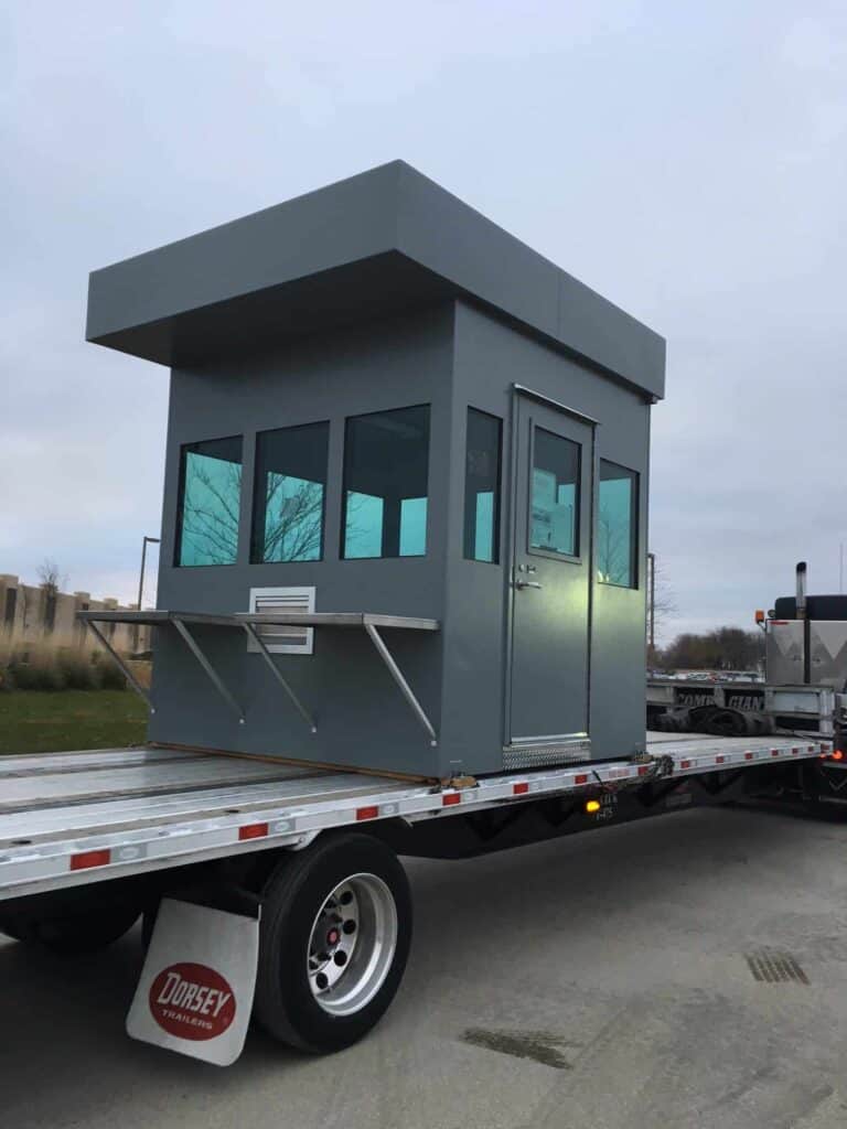 Guard/Security Booth 37 A portable gray security booth with access control and multiple windows is mounted on a flatbed trailer parked outdoors under a cloudy sky.
