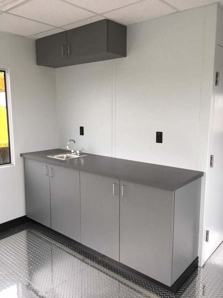 Guard/Security Booth 34 A clean, modern kitchenette with a stainless steel sink set into gray cabinetry, overhead cabinets, and a glossy tiled floor, illuminated by natural light from a small window near security booths.
