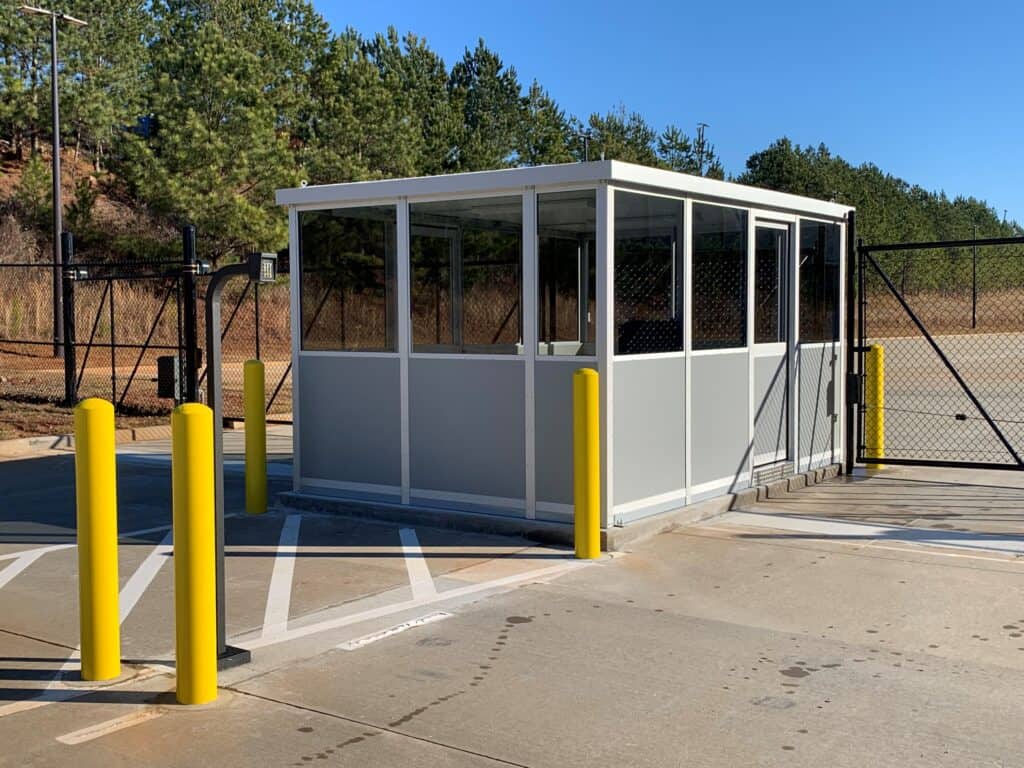 Guard/Security Booth 1 A small, modern guard shack with glass windows, positioned at the entrance of a parking area, surrounded by yellow bollards and a metal fence, under a clear blue sky.