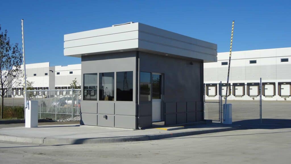 Guard/Security Booth 30 A small, modern portable security booth with large windows situated at the entrance of a secured industrial area, featuring white barriers and fencing.
