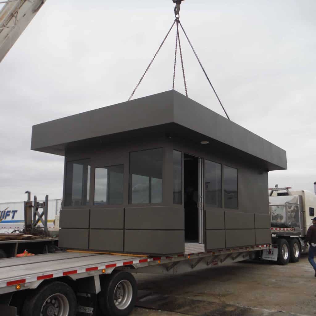 A prefabricated guard shack being lifted by a crane from a flatbed trailer at an industrial site.