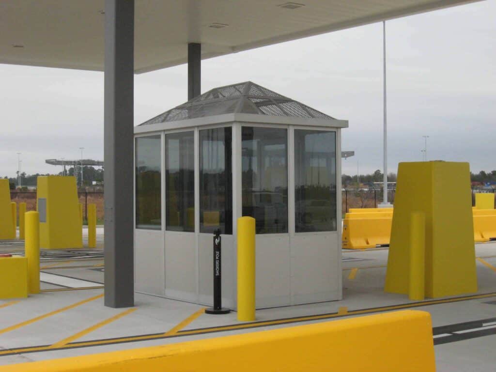 Guard/Security Booth 46 A small, modern glass security booth with a gray roof, located at a parking lot entrance, surrounded by bright yellow barriers under a cloudy sky.