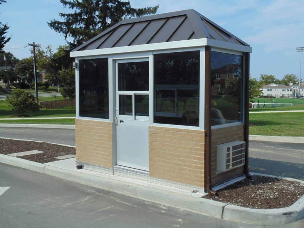 Guard/Security Booth 27 A small, modern security booth with glass walls and a dark metal roof, situated on a newly paved area with surrounding grass and trees in a suburban setting.