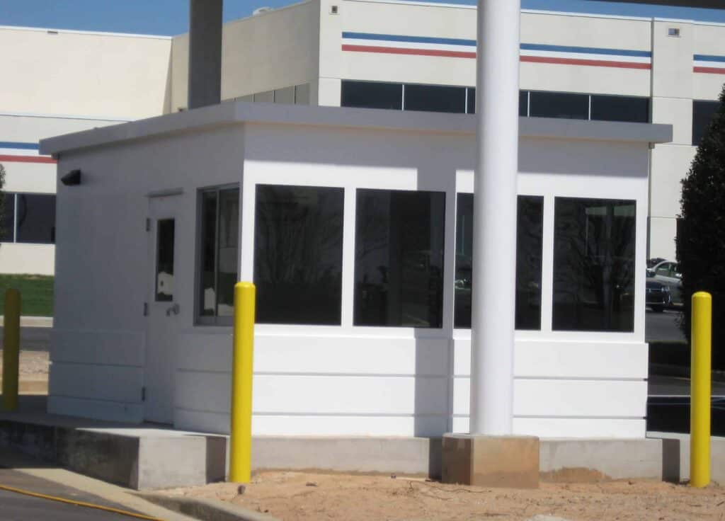 Guard/Security Booth 48 A small, white guard shack with large windows, surrounded by yellow bollards, located in a parking lot with a large building in the background.