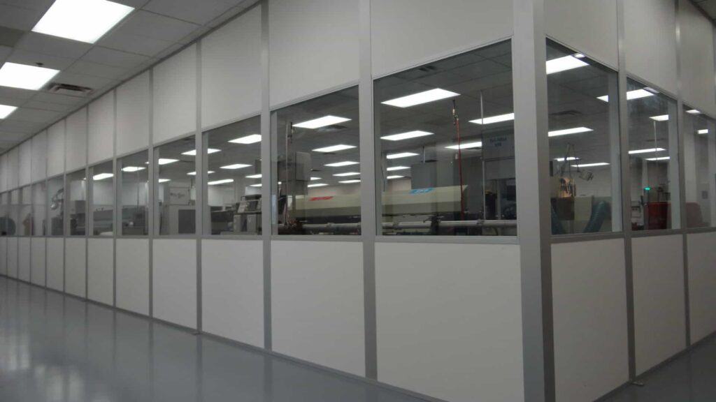 A view through large glass windows into a clean, well-lit industrial workspace with rows of indoor equipment enclosures and machinery, featuring a minimalistic and modern design.