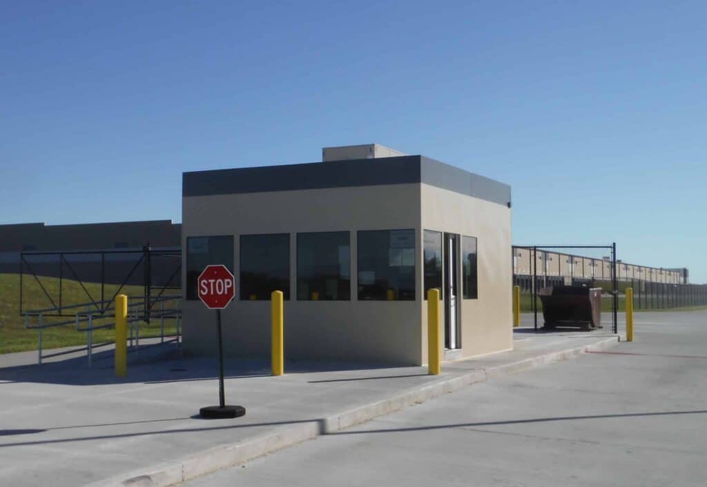 Guard/Security Booth 32 A small, modern access control booth at the entrance of a facility with a stop sign and yellow bollards under a clear blue sky.