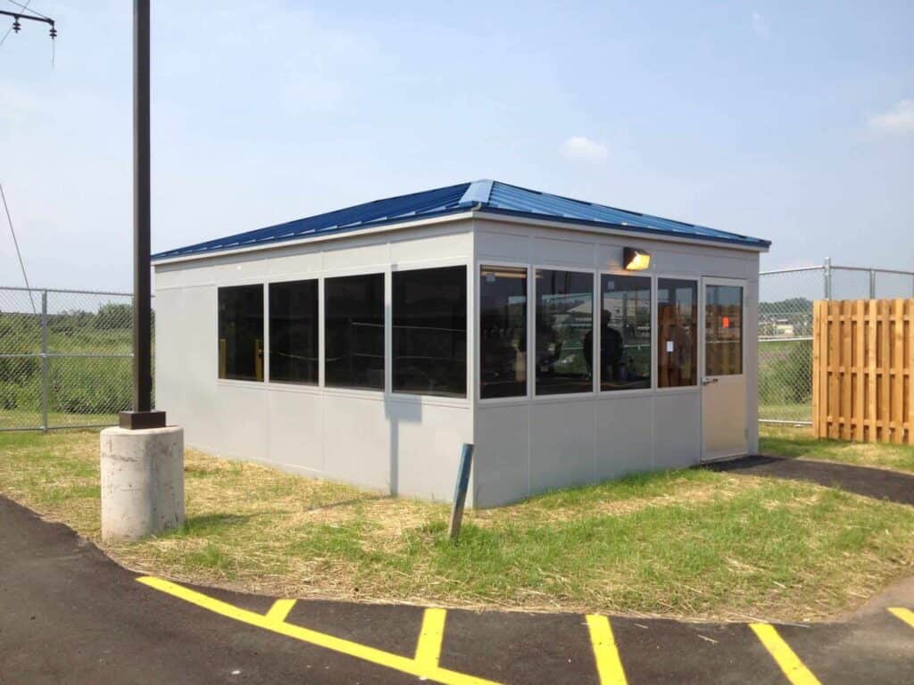 Guard/Security Booth 29 A small, modern security booth with large windows and a blue roof, situated on a grassy area next to a yellow-lined pavement and a wooden fence.