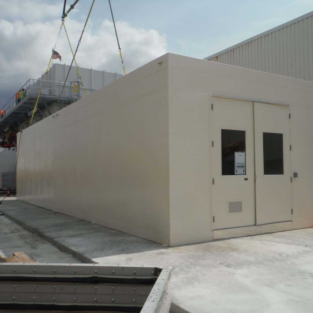 Motor Control Center Enclosure 3 A large, beige MCC enclosure with a flat roof being installed next to a factory, featuring a single door and several windows, under a cloudy sky. A crane is visible in the background.