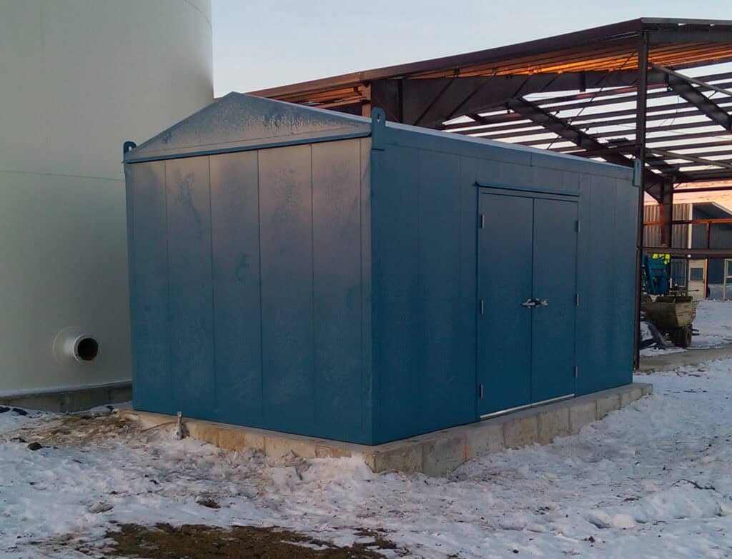 Motor Control Center Enclosure 2 A blue utility building with a sloped roof, located next to a large storage tank and under a partially constructed motor control center (MCC), surrounded by snow.
