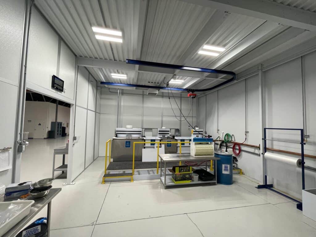 A clean and organized industrial workspace with white walls and ceiling, featuring various workstations, shelves, and an overhead crane system. Bright lighting illuminates the area, which includes protective enclosures around critical machinery
