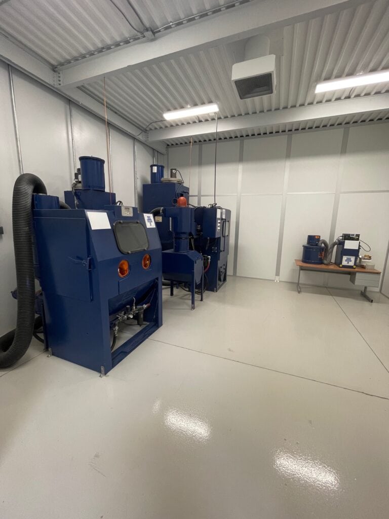 Interior of a clean industrial workshop with two large blue machines equipped with noise reduction enclosures and various equipment, under bright lighting in a room with white walls and gray flooring.