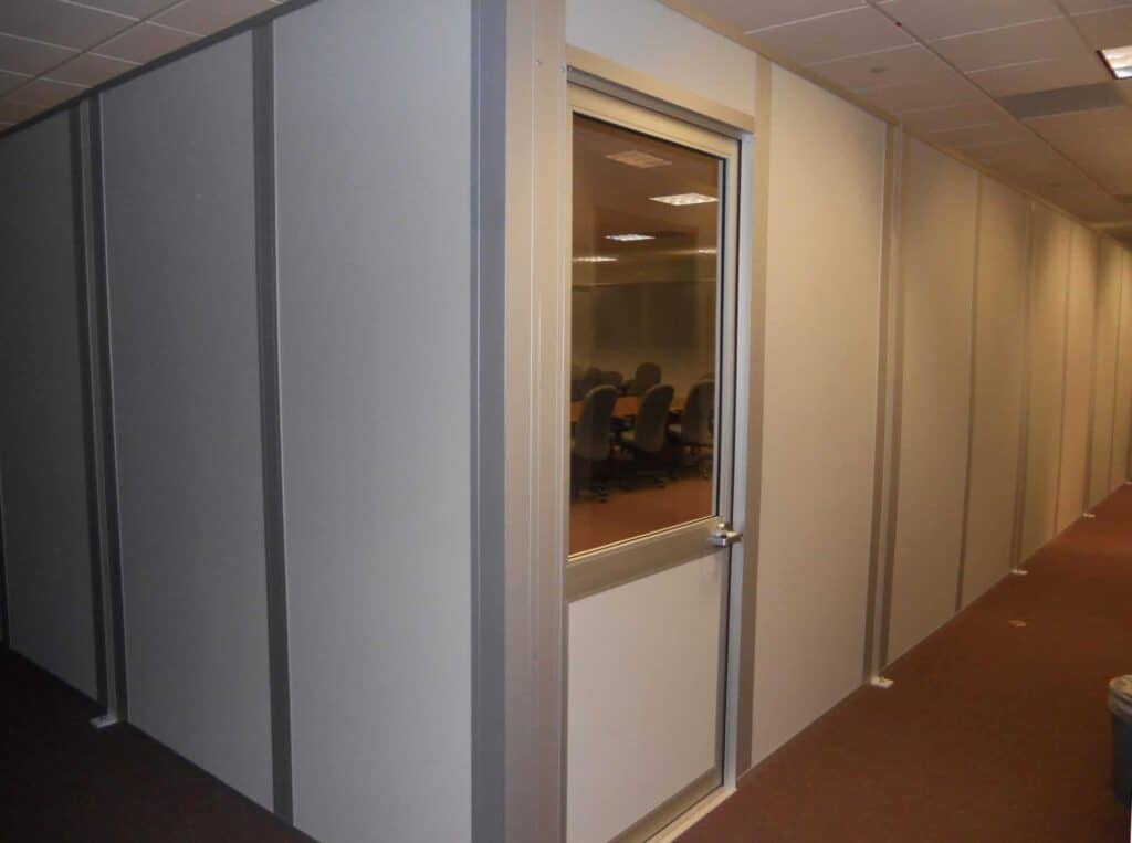 A corridor with gray modular partitions and a glass door leading into a well-lit office room with chairs and a table visible through the window.