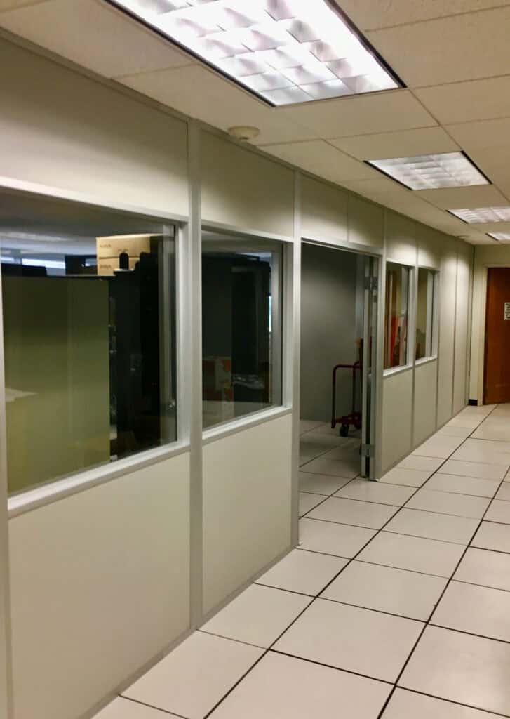A corridor with modular office walls on one side and a white tiled floor. Fluorescent lighting illuminates the hallway.