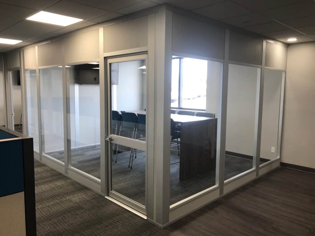 Modern office meeting room enclosed by glass wall partitions with a door, featuring a conference table and chairs inside, situated in a well-lit space with carpeted flooring.
