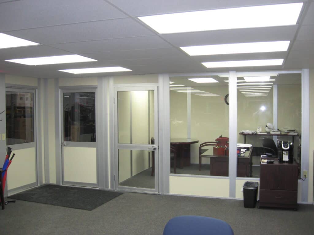 Modern office space with bright fluorescent lighting. The area features glass office partitions, a desk with a computer, chairs, and visible office equipment. A clean and organized environment.