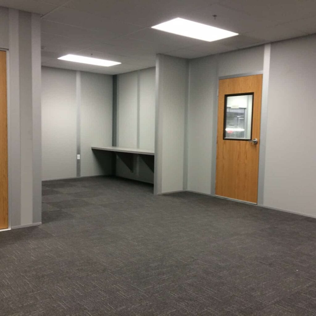 A modern office space with gray carpet, white walls, office partitions, a wooden door with a glass window, and a built-in gray counter along the left wall.