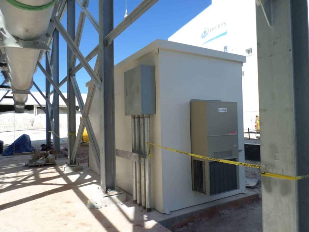 An electrical utility room with a closed metal door is surrounded by outdoor equipment enclosures and concrete columns at an industrial site. Caution tape is visible around the site.
