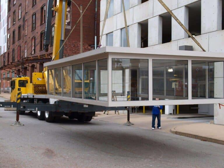 A construction worker in a blue jacket observes a large prefab shelter being transported on a flatbed trailer near a building under construction, with a yellow crane and urban backdrop.