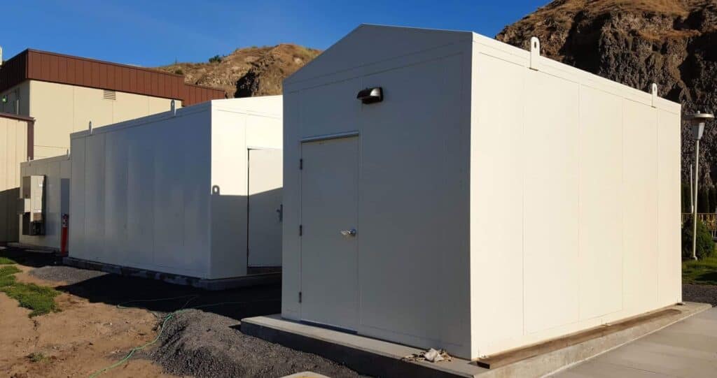 Three large white outdoor equipment enclosures with a door and a security camera mounted on one, situated on gravel ground near a rocky hillside under a clear sky.