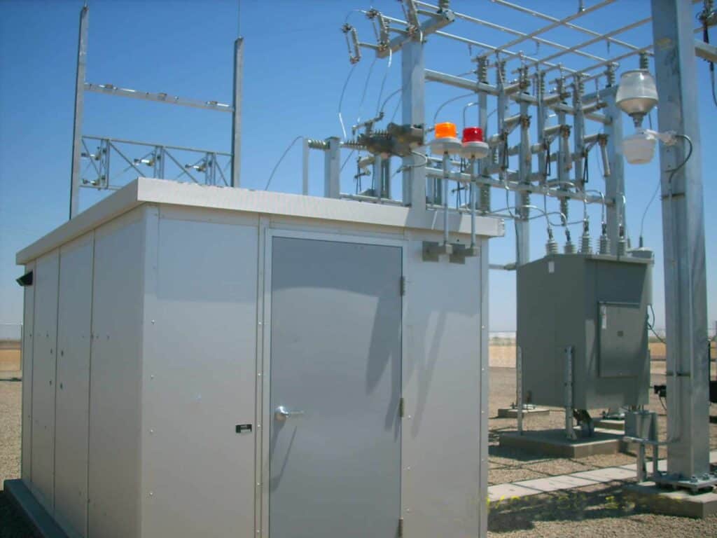 An electrical substation in a desert environment, featuring a control building and various outdoor equipment including transformers and disconnect switches under a clear blue sky.