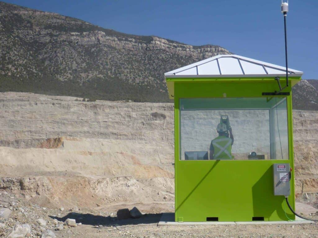 A green security enclosure stationed at a quarry with rocky, barren mountains in the background under a clear blue sky. A mannequin sits inside the booth appearing to monitor the area.