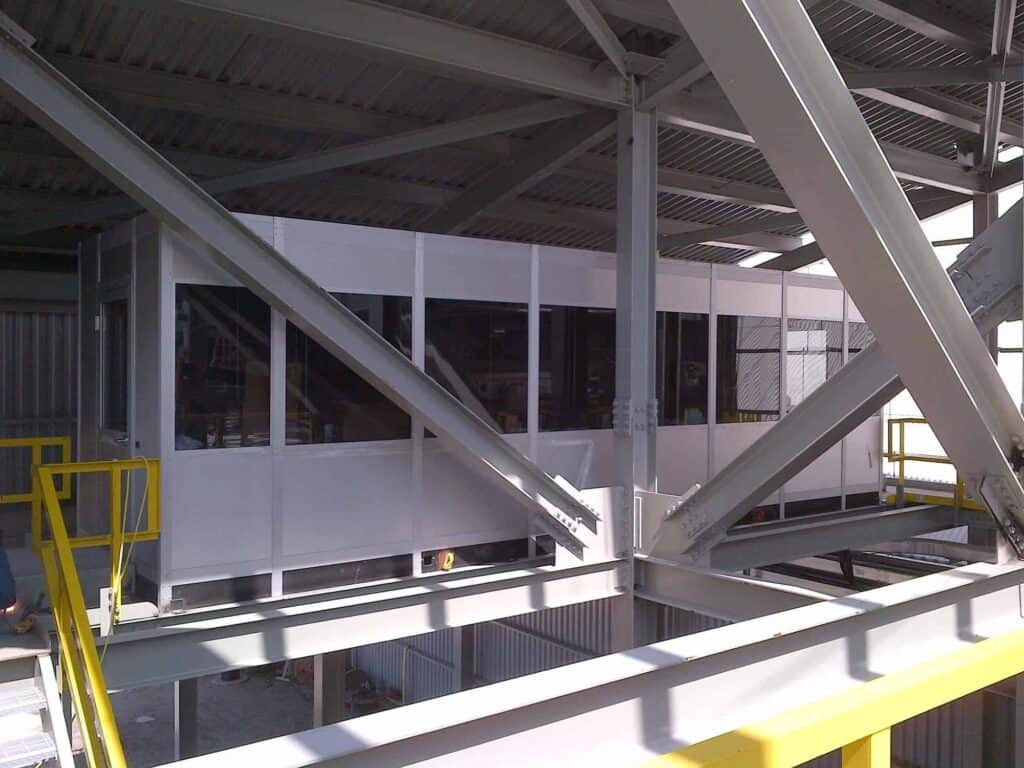 Industrial setting with large steel beams supporting the structure and outdoor equipment enclosures in the background. Visible metallic and yellow railings enhance safety.