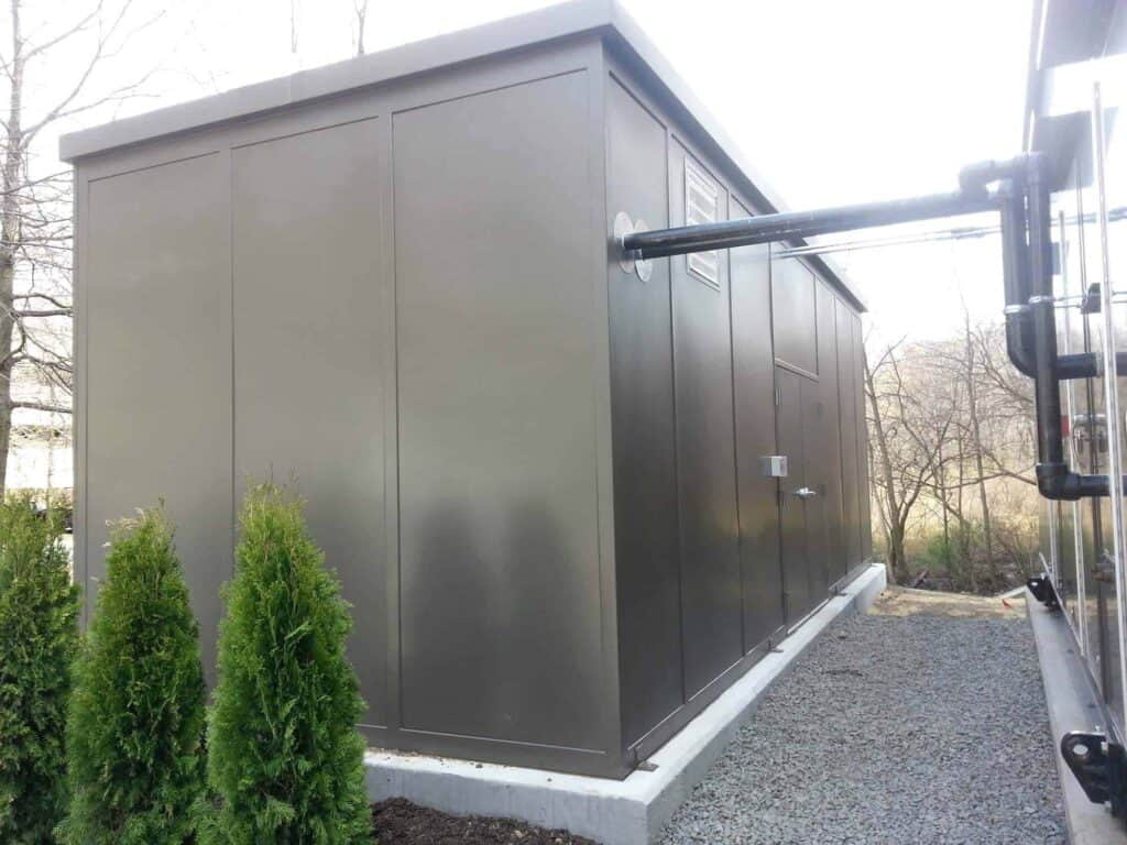 A modern, gray outdoor equipment enclosure with a flat roof, located in a garden with small shrubs and crushed stone pathway beside a metal fence.