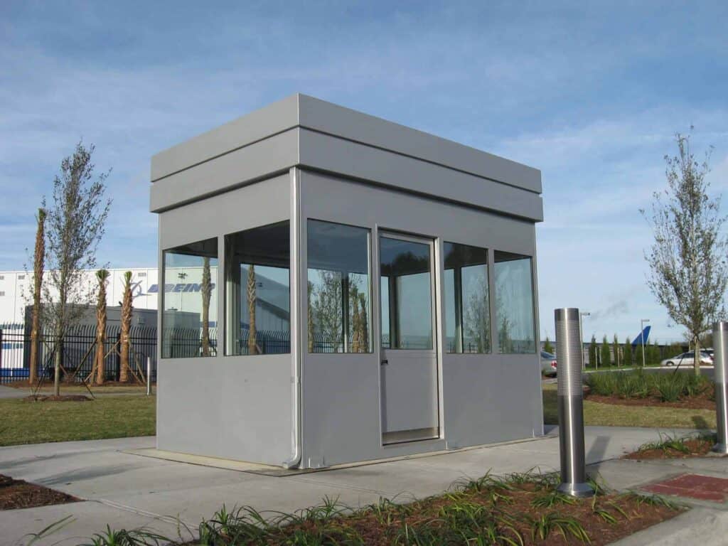 Guard/Security Booth 3 A small, modern modular security booth with glass walls and a gray flat roof, located on a concrete path beside some greenery and young trees under blue skies.